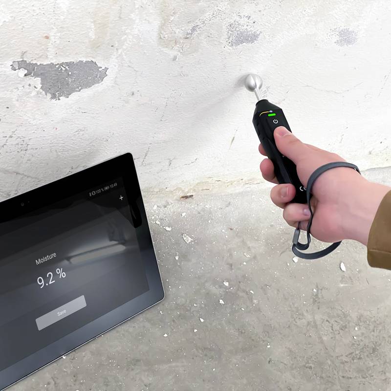 Eine Hand hält ein Feuchtigkeitsmessgerät an einer Wand. Ein daneben liegendes Tablet zeigt '9,2 %' an.
