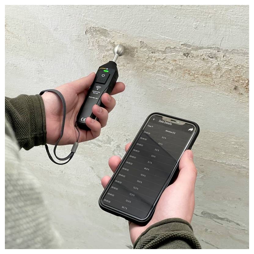 Eine Person hält ein Feuchtemessgerät gegen eine feuchte, abblätternde Wand, während sie die Feuchtemesswerte in einer Smartphone-App betrachtet.