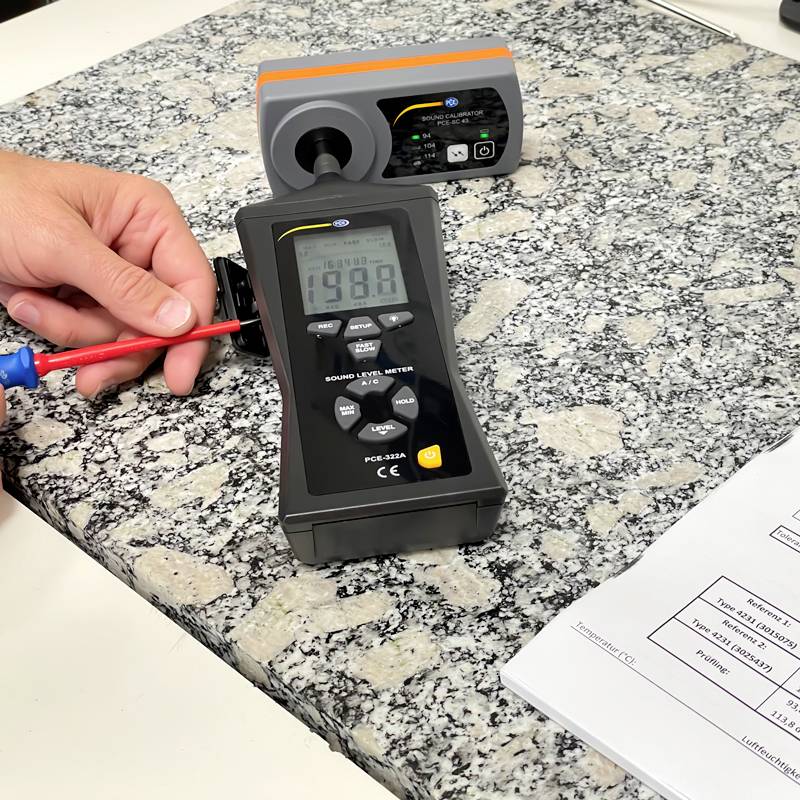 Hand hält einen Spannungsmesser, der 198 V auf einem Steinfußboden anzeigt, neben einem schwarzen und grauen Prüfgerät.