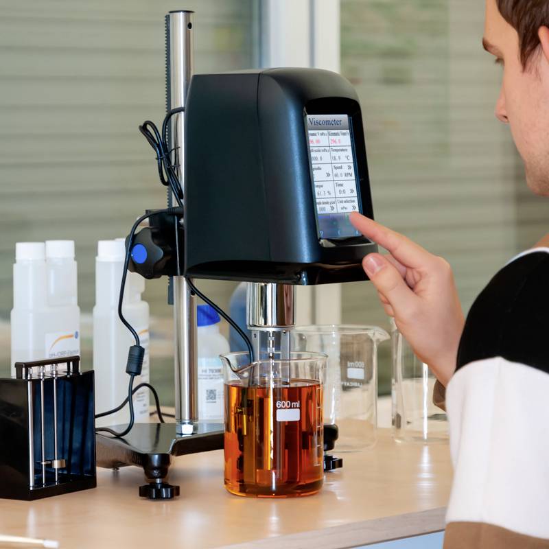 Eine Person bedient ein Viskosimeter in einem Labor. Ein Becherglas mit Flüssigkeit steht auf dem Gerät. Im Hintergrund sind Laborutensilien.