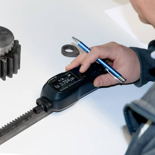 Eine Person misst mit einem digitalen Messgerät den Abstand einer geriffelten Metallstange. Daneben liegt ein Zahnrad.