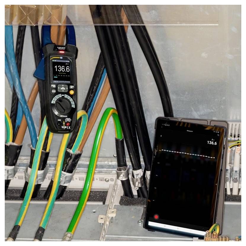 Elektrisches Schaltfeld mit unter Spannung stehenden Leitungen und Stromzange mit Anzeige von 136,6 A; ein Tablet spiegelt die Stromstärkeanzeige zur Überwachung.