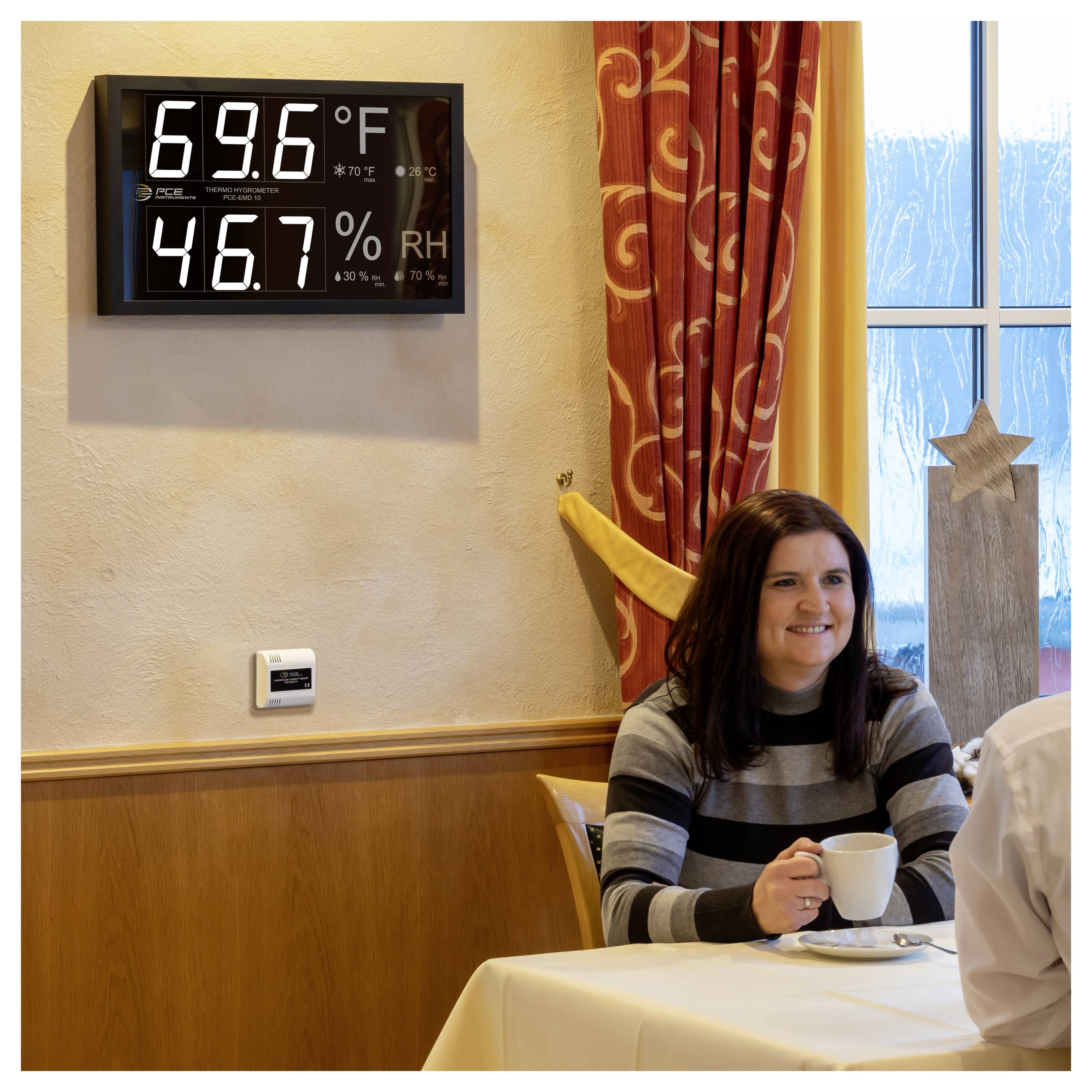 Eine Frau, die an einem Tisch in einem Café lächelt, hält eine Tasse in der Nähe eines Fensters; eine Wandanzeige zeigt 69,6°F und 46,7% Luftfeuchtigkeit.