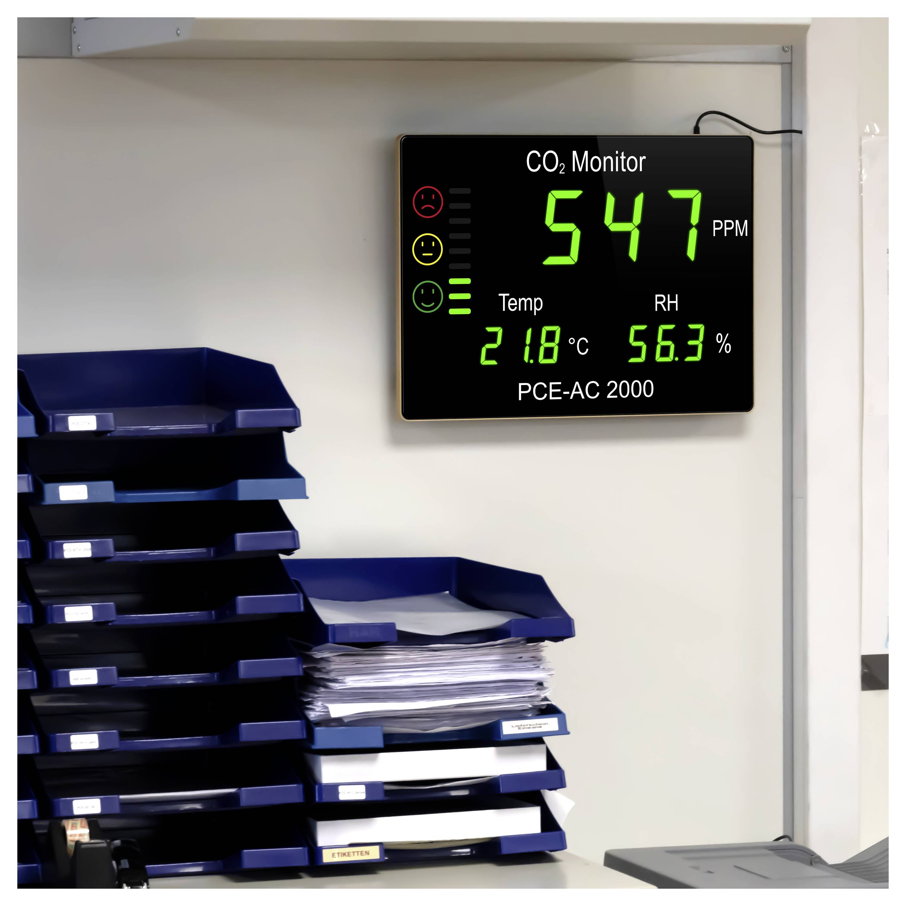 CO2-Monitor an der Wand zeigt 547 ppm, 21,8°C und 56,3% Luftfeuchtigkeit über gestapelten Dokumentablage­fächern in einem Büro.