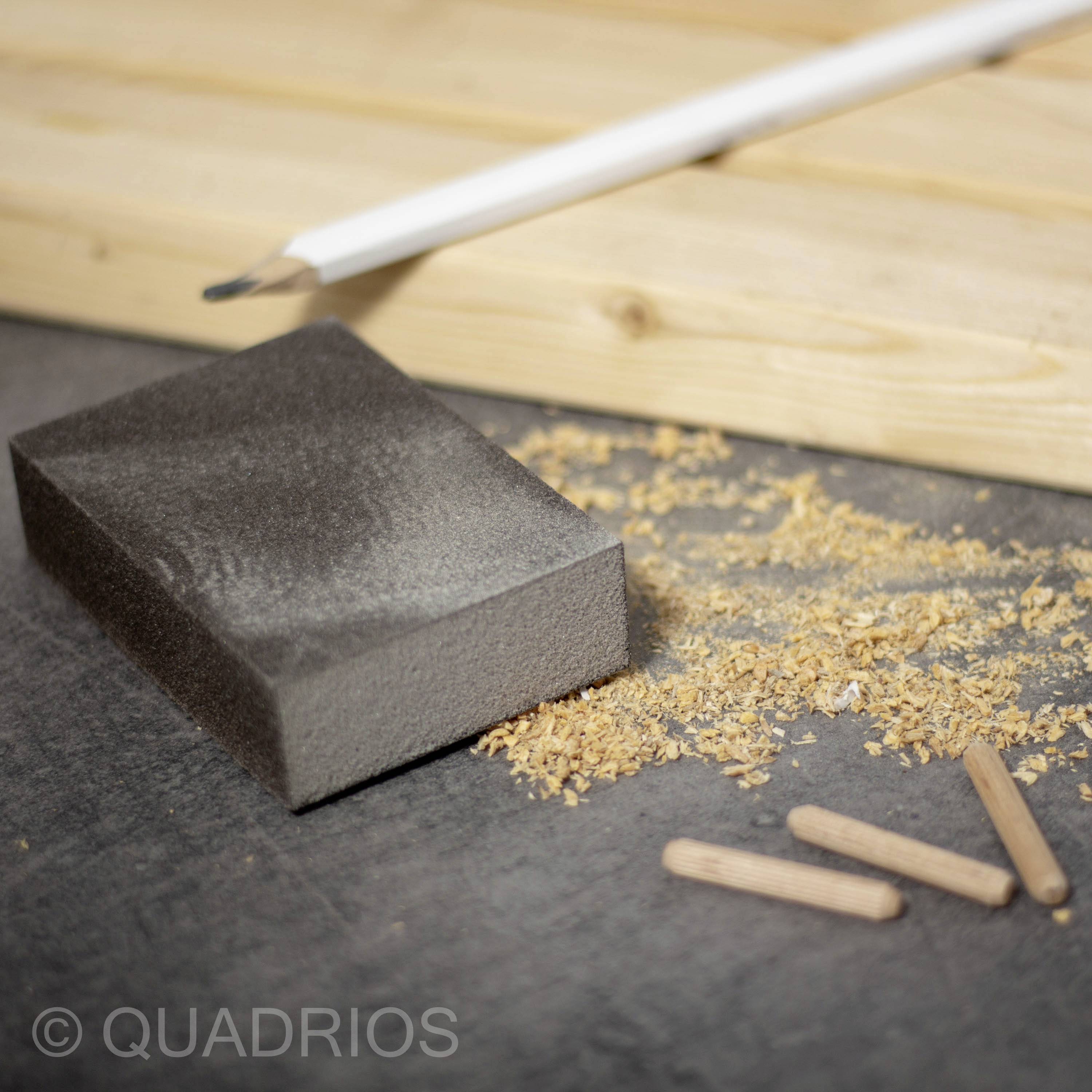 Schleifblock und Sägespäne auf einer Werkbank mit Holzdübeln im Vordergrund, Bleistift liegt quer im Hintergrund.