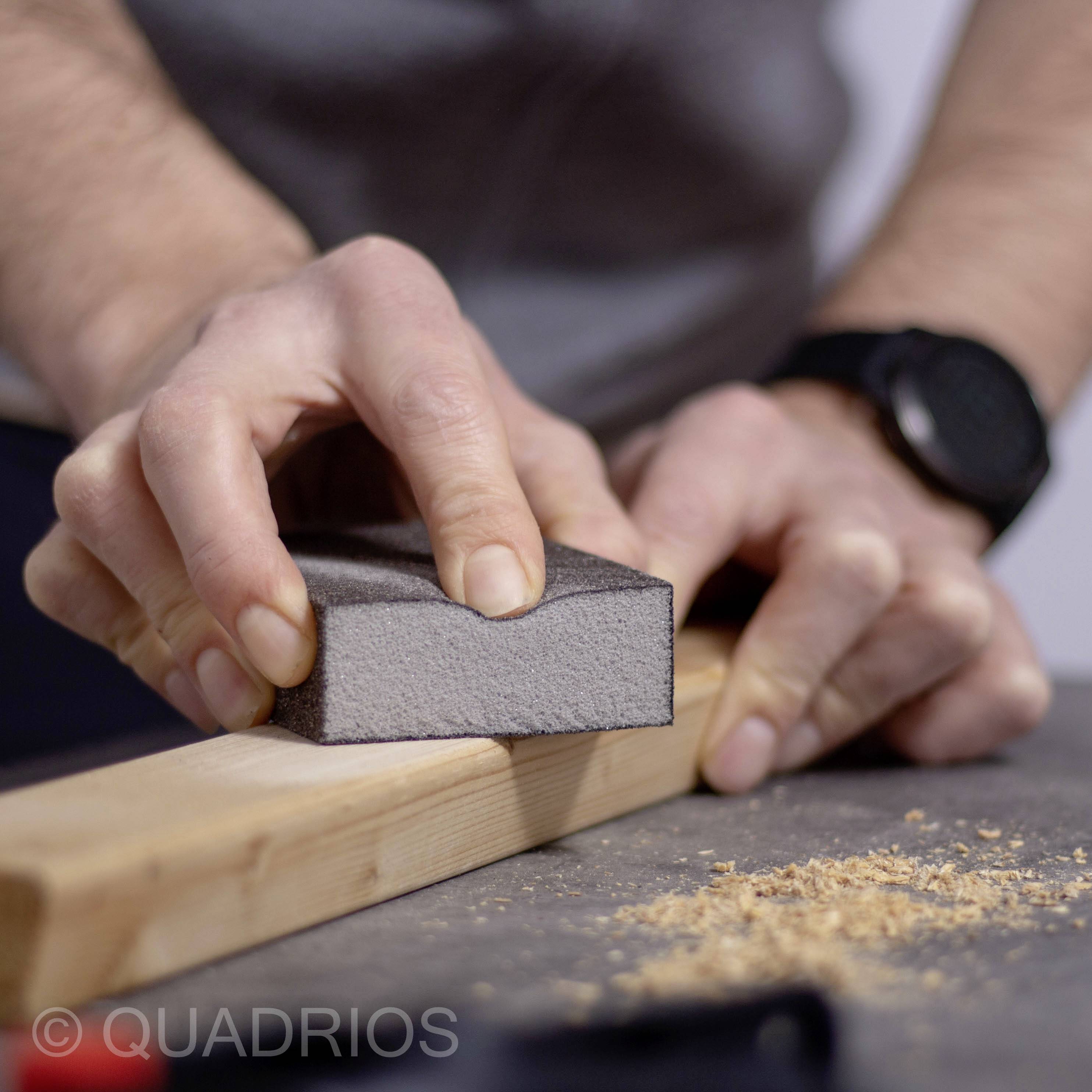 Eine Person glättet ein Holzbrett mit einem Schleifblock, Holzspäne liegen daneben. Deutlicher Fokus auf Präzision und Handwerk.