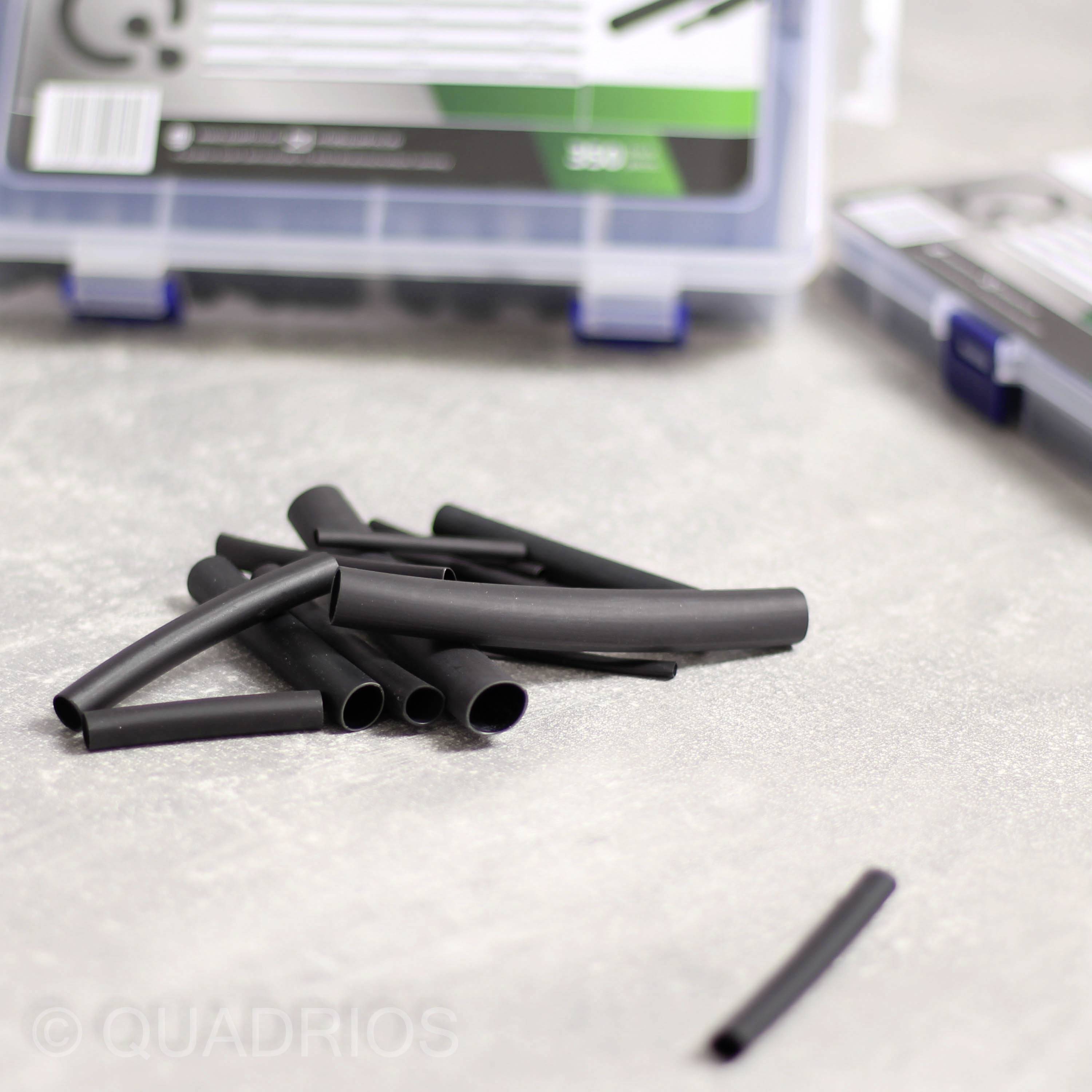 Ein Stapel schwarzer Schrumpfschläuche liegt auf einer grauen Oberfläche, im Hintergrund sind halb geöffnete Plastikboxen sichtbar.