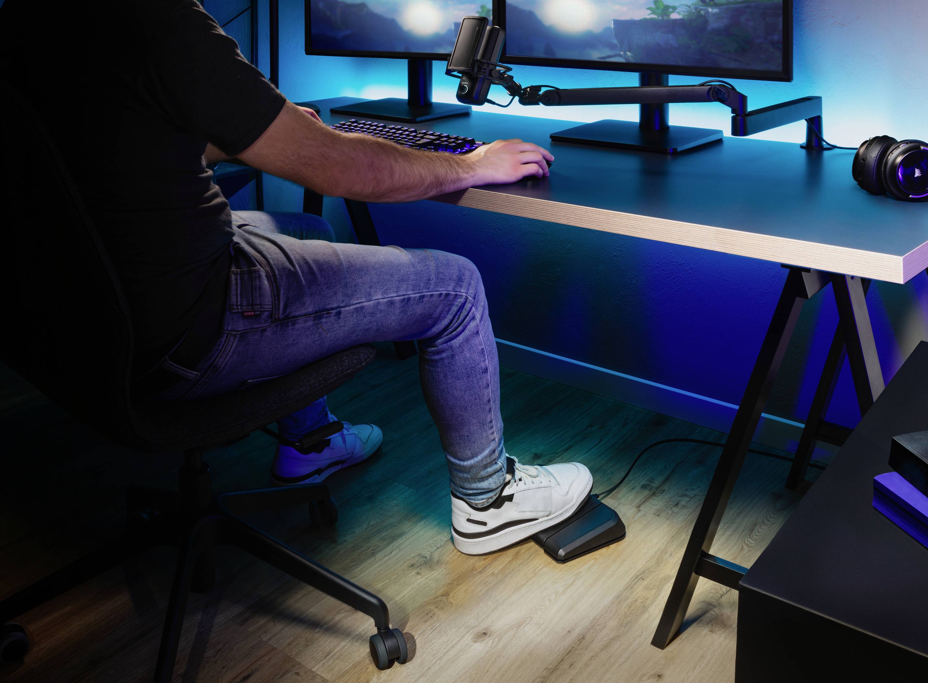 Eine Person sitzt an einem Schreibtisch mit Monitor und Mikrofon, trägt Kopfhörer und bedient mit dem Fuß ein Pedal unter dem Tisch.