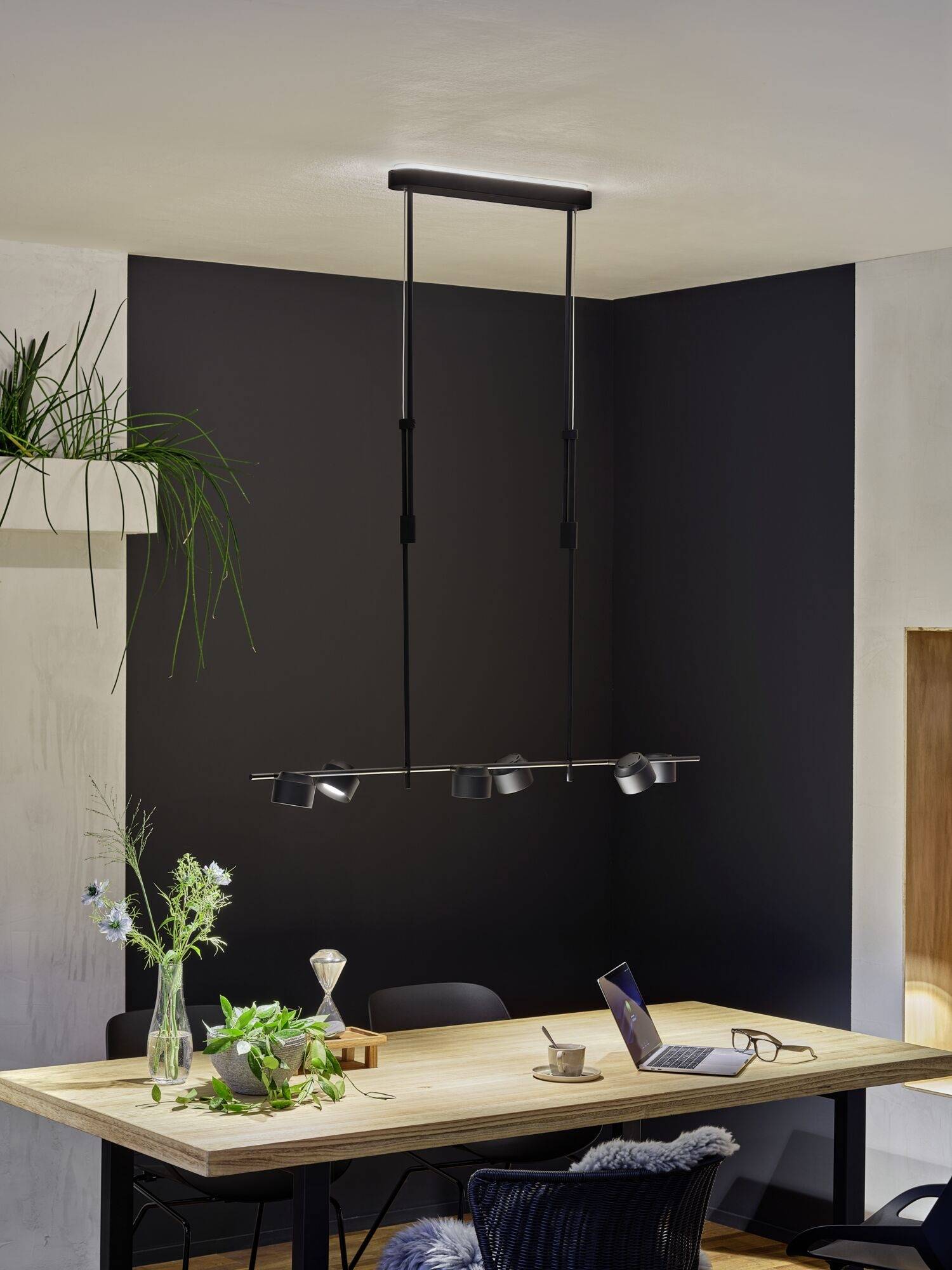 Ein moderner Esstisch mit einer hängenden, minimalistischen Lampe darüber. Auf dem Tisch stehen Pflanzen, Bücher und ein Laptop.