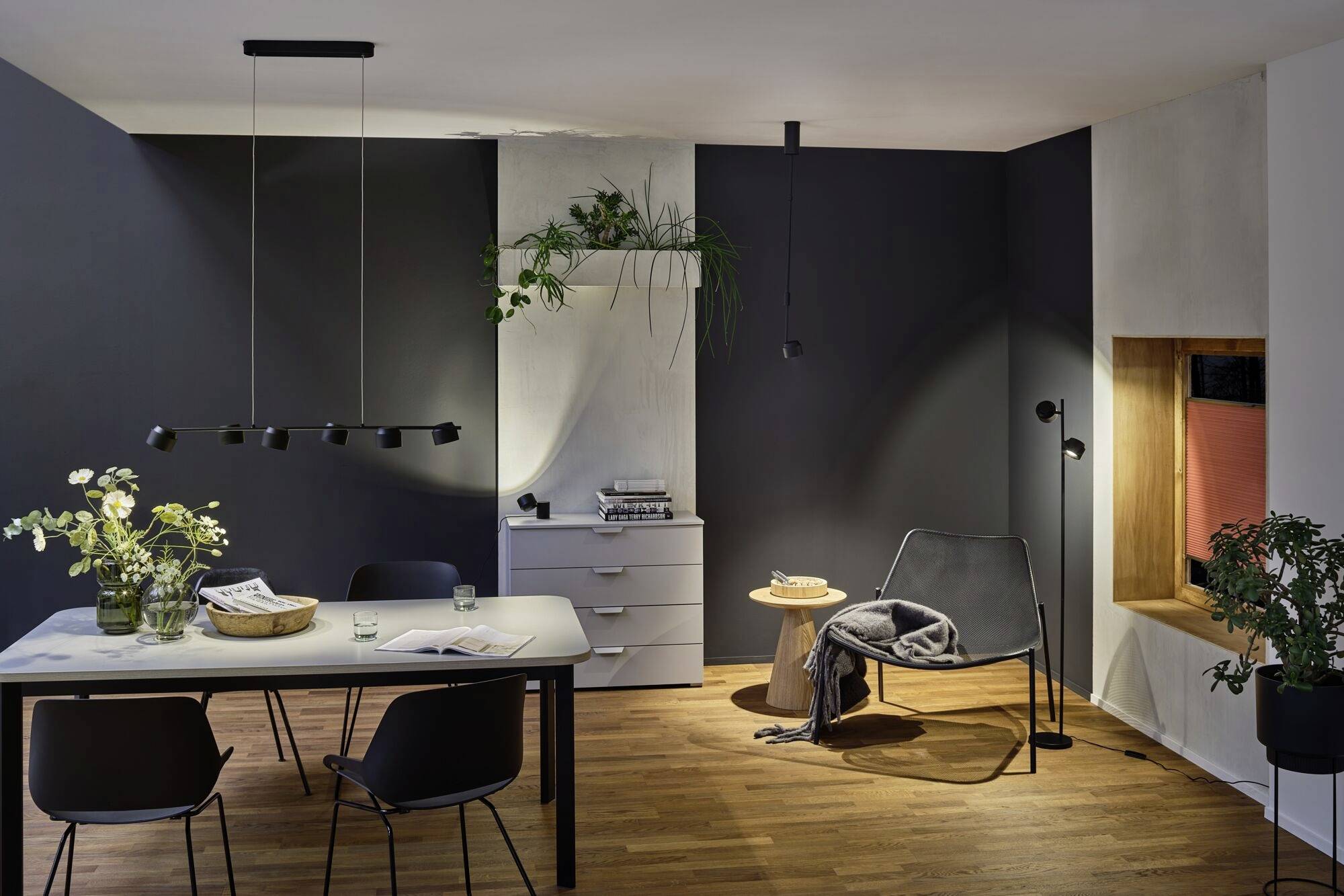 Ein modernes Wohnzimmer mit dunklen Wänden, Holzboden, einem Esstisch mit vier Stühlen, einer gemütlichen Ecke mit Sessel und Pflanzen.