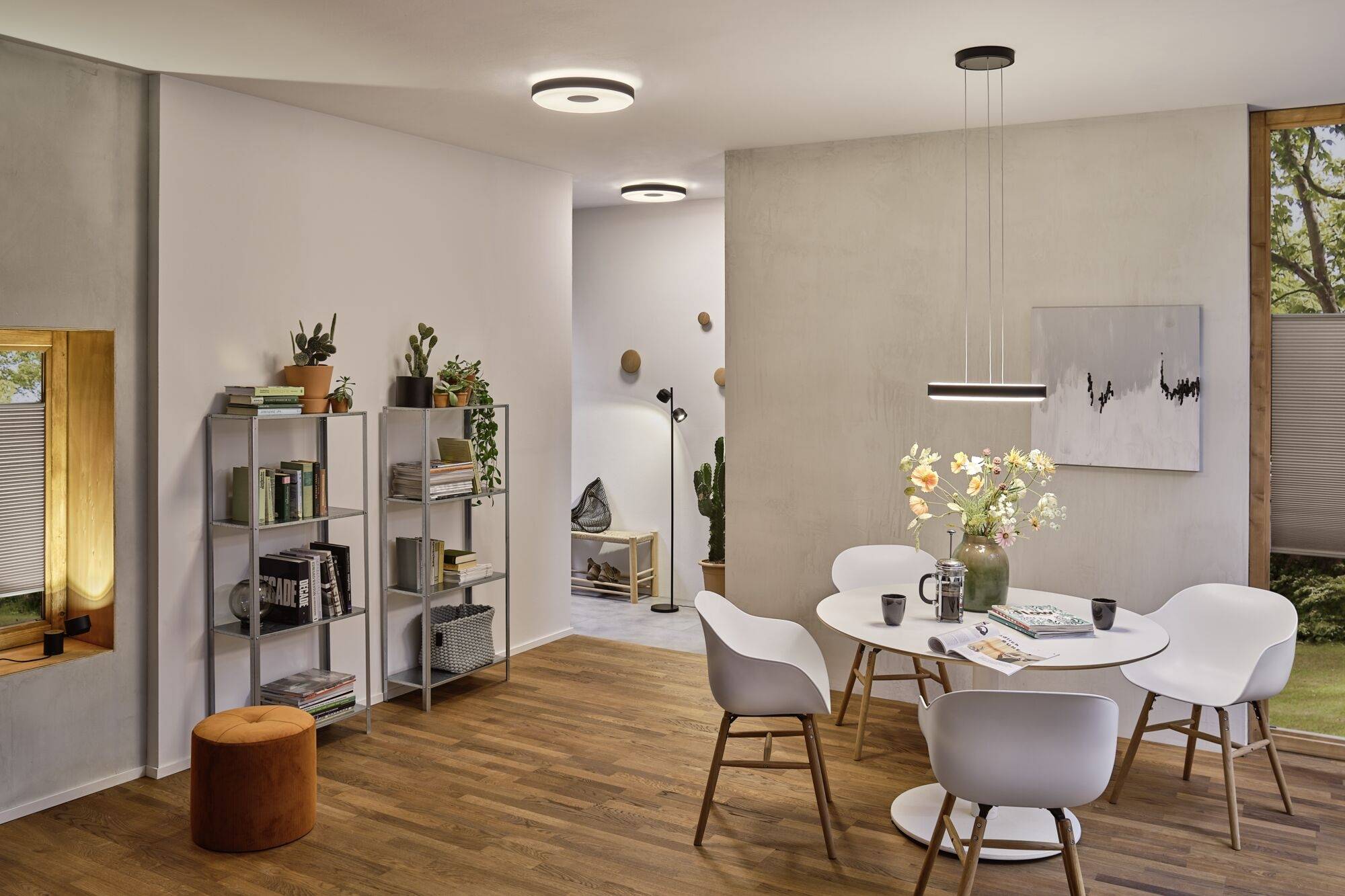 Essbereich mit rundem Tisch, fünf weißen Stühlen, hellen Wänden und Holzfußboden. Links Regal, rechts Bild und Pflanzen auf Tisch.