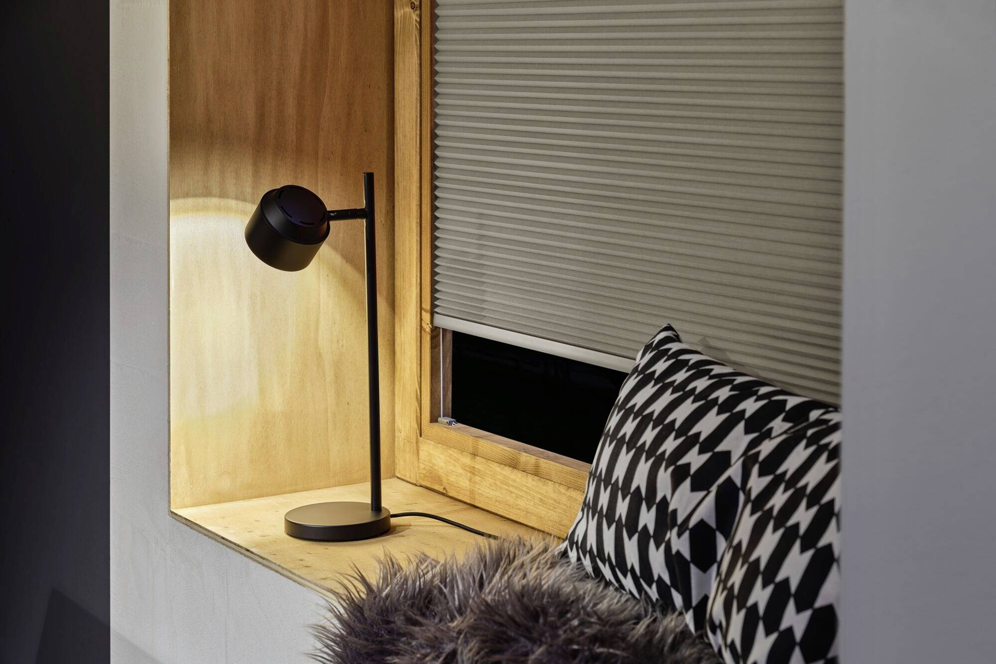 Gemütliche Leseecke mit einer leuchtenden Tischlampe, schwarz-weiß gemustertem Kissen und Fellkissen auf einer Fensterbank.