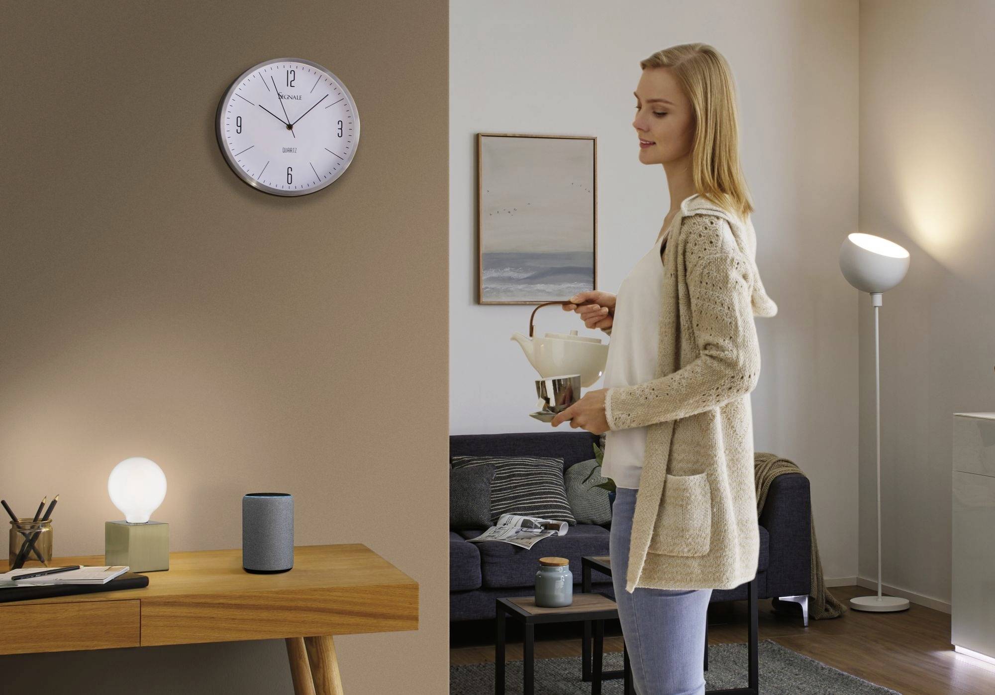 Eine Frau steht in einem Wohnzimmer, hält einen Kessel und gießt Tee ein. Im Hintergrund eine Uhr an der Wand und Möbel.