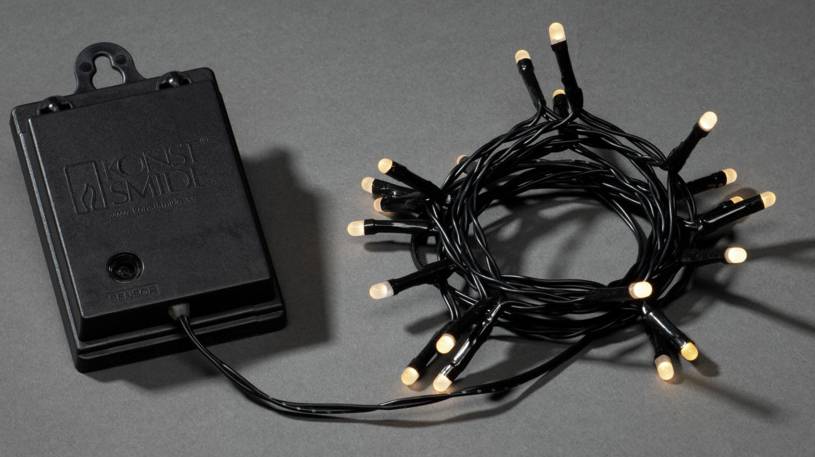Ein schwarzer Batteriekasten und eine zusammengewickelte Lichterkette mit leuchtenden, kleinen Glühbirnen auf einem grauen Hintergrund.