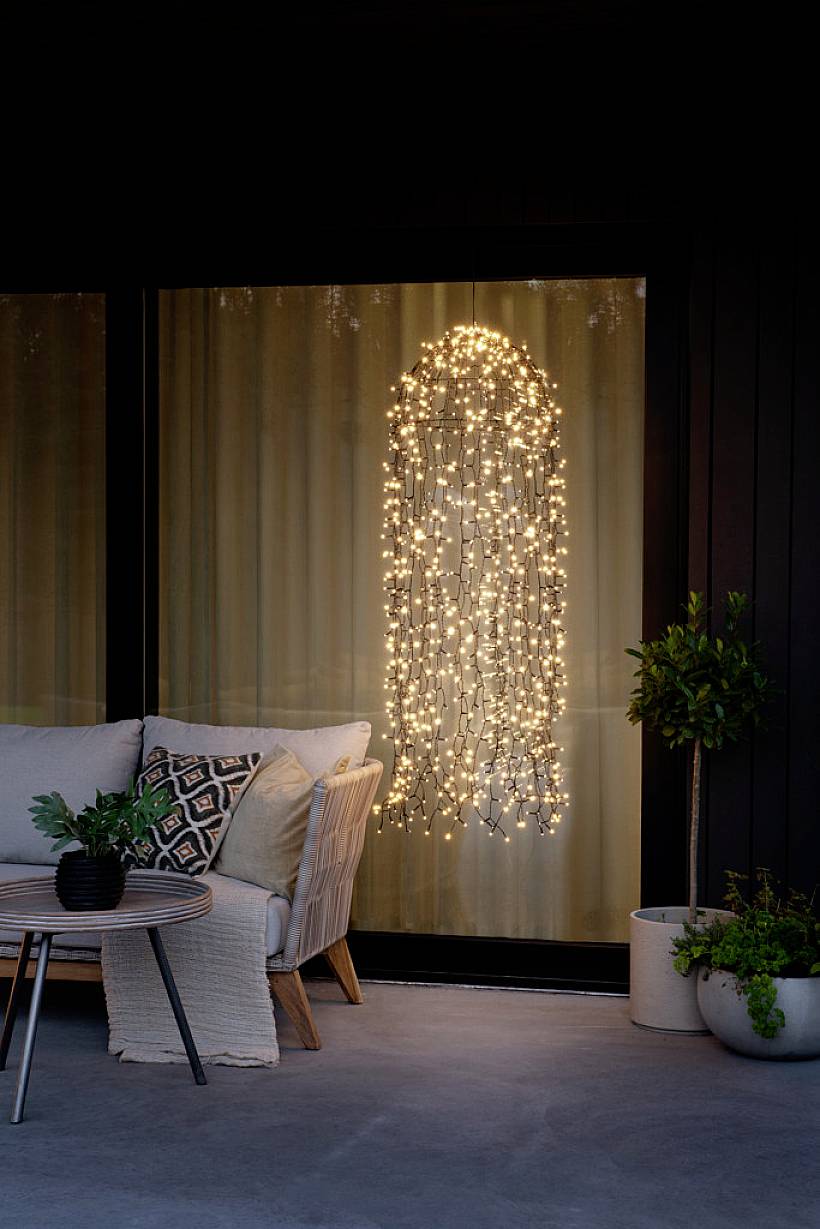 Gemütliche Terrassenszene mit beleuchtetem Vorhang aus Lichterketten, Sitzecke mit Kissen und Pflanzen vor einem Fenster.