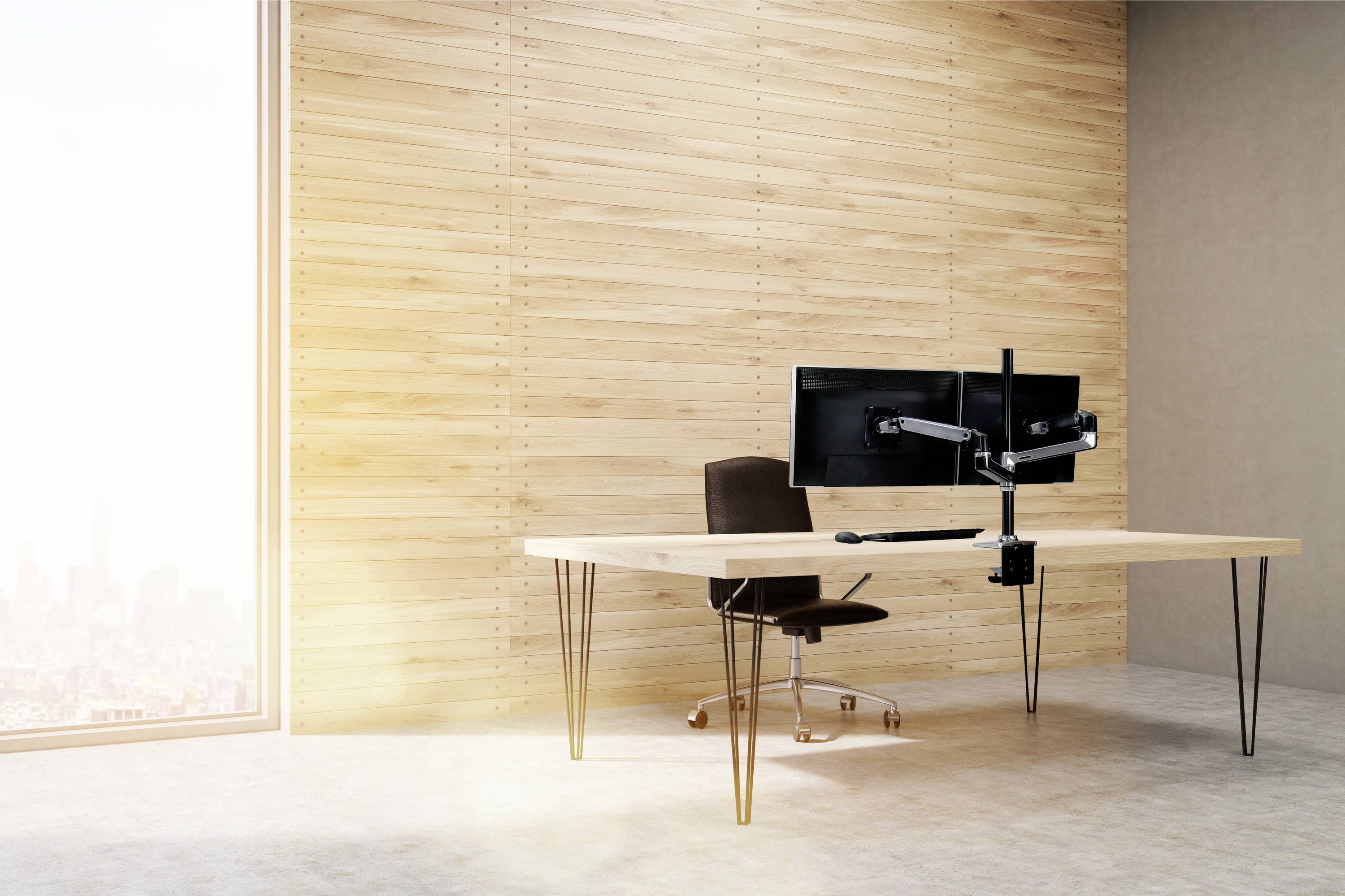 Moderner Schreibtisch mit zwei Monitoren auf einem Monitorarm und einem schwarzen Stuhl. Der Raum hat Holzwände und einen großen Fensterblick.