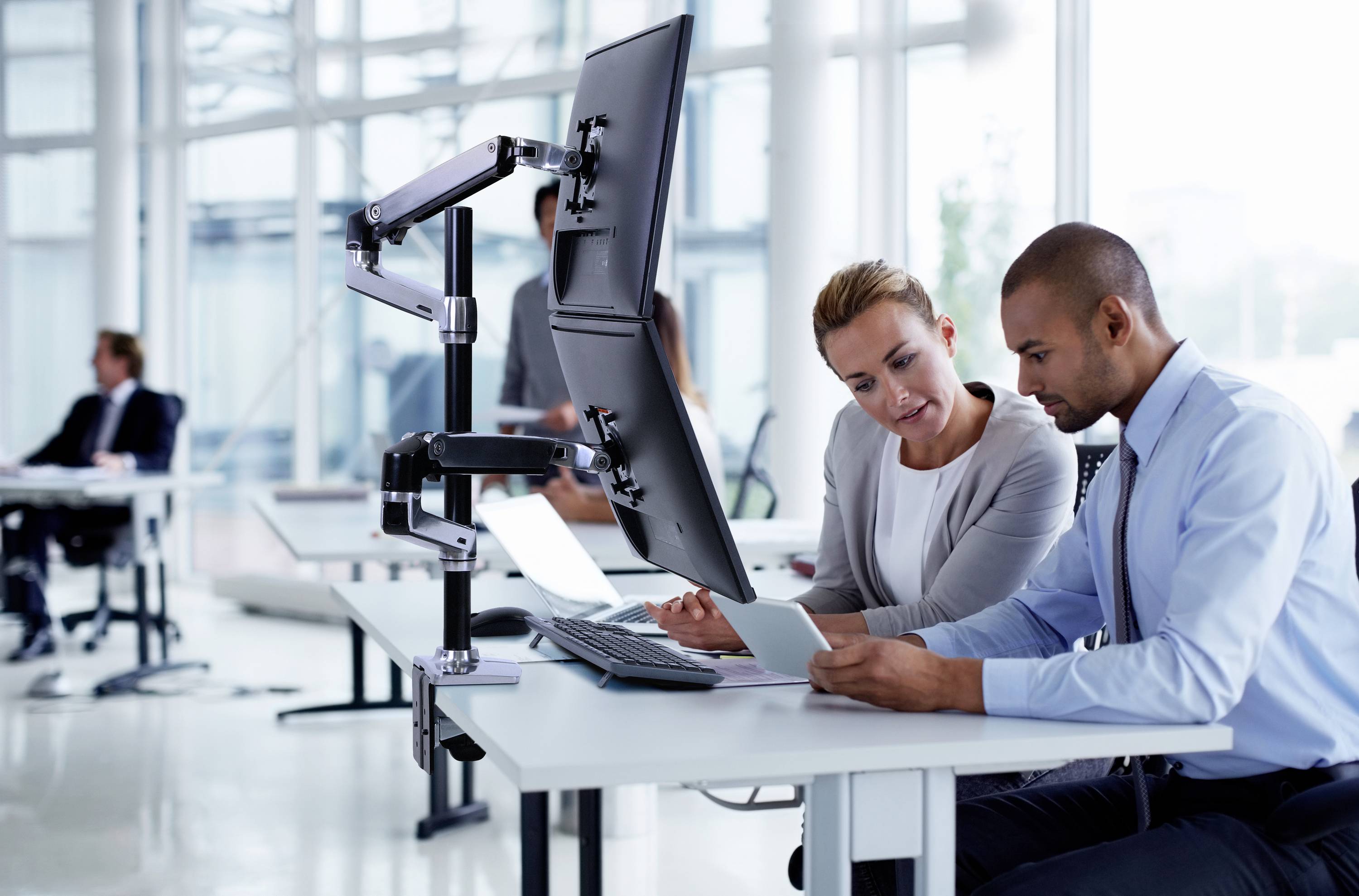 Ein Mann und eine Frau sitzen an einem Schreibtisch in einem modernen Büro und schauen auf ein Tablet. Hinter ihnen sind Arbeitskollegen.