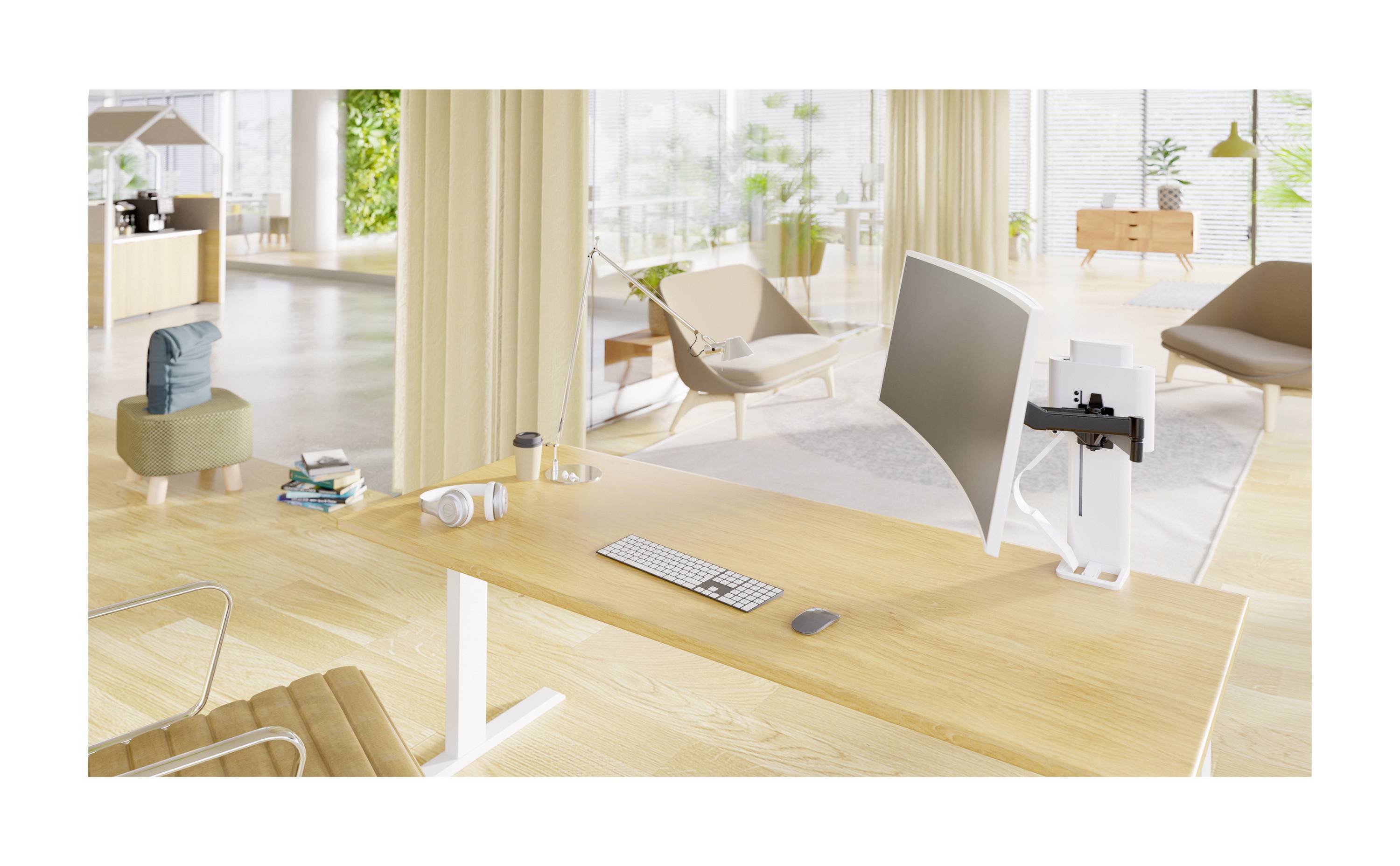 Moderner Büroraum mit einem Schreibtisch, darauf ein Monitor, Tastatur und Maus. Im Hintergrund sind Sitzbereiche und große Pflanzen sichtbar.