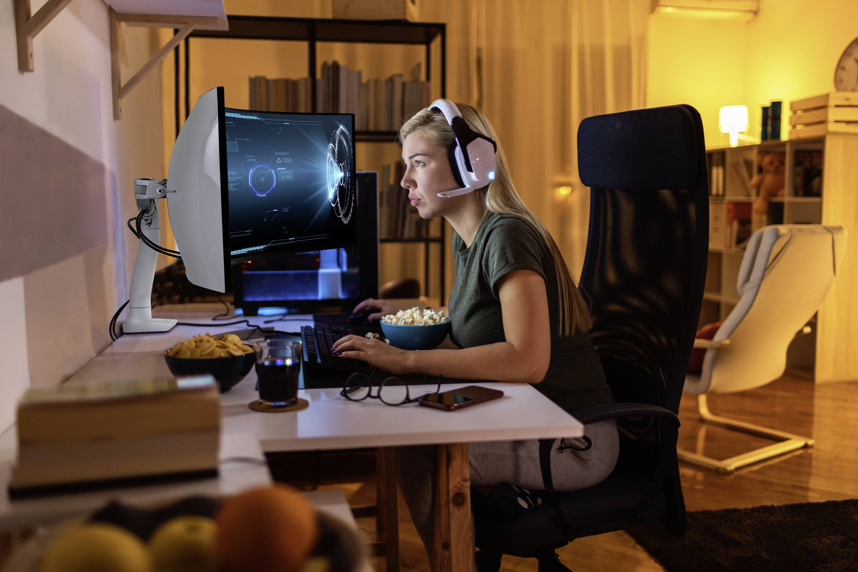Eine Person trägt ein Headset und spielt abends ein Videospiel am Computer. Auf dem Tisch stehen Snacks und ein Getränk.