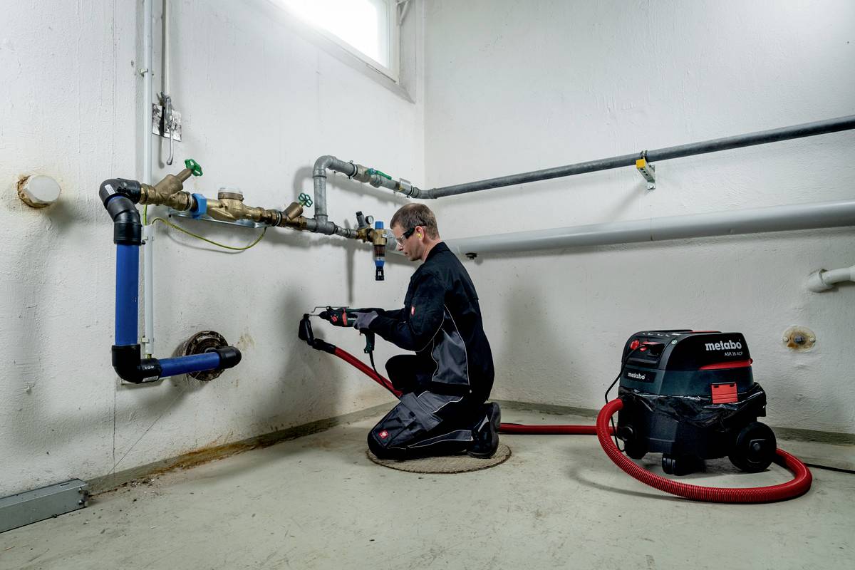 Eine Person in Arbeitskleidung bedient ein Gerät in einem Technikraum. Ein Staubsauger ist neben ihnen mit einem Schlauch verbunden.