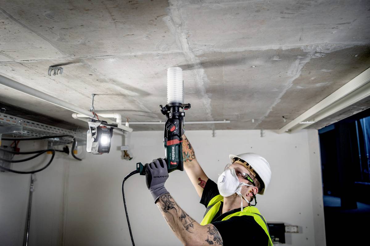 Ein Bauarbeiter in Sicherheitskleidung und Atemschutzmaske bohrt mit einer elektrischen Bohrmaschine in eine Betondecke.