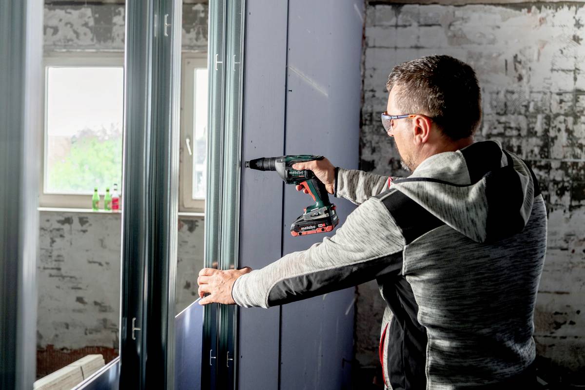 Eine Person befestigt mit einem Akkubohrer Metallrahmen an einer Wand in einem renovierten Raum mit freigelegtem Mauerwerk und Fenstern.