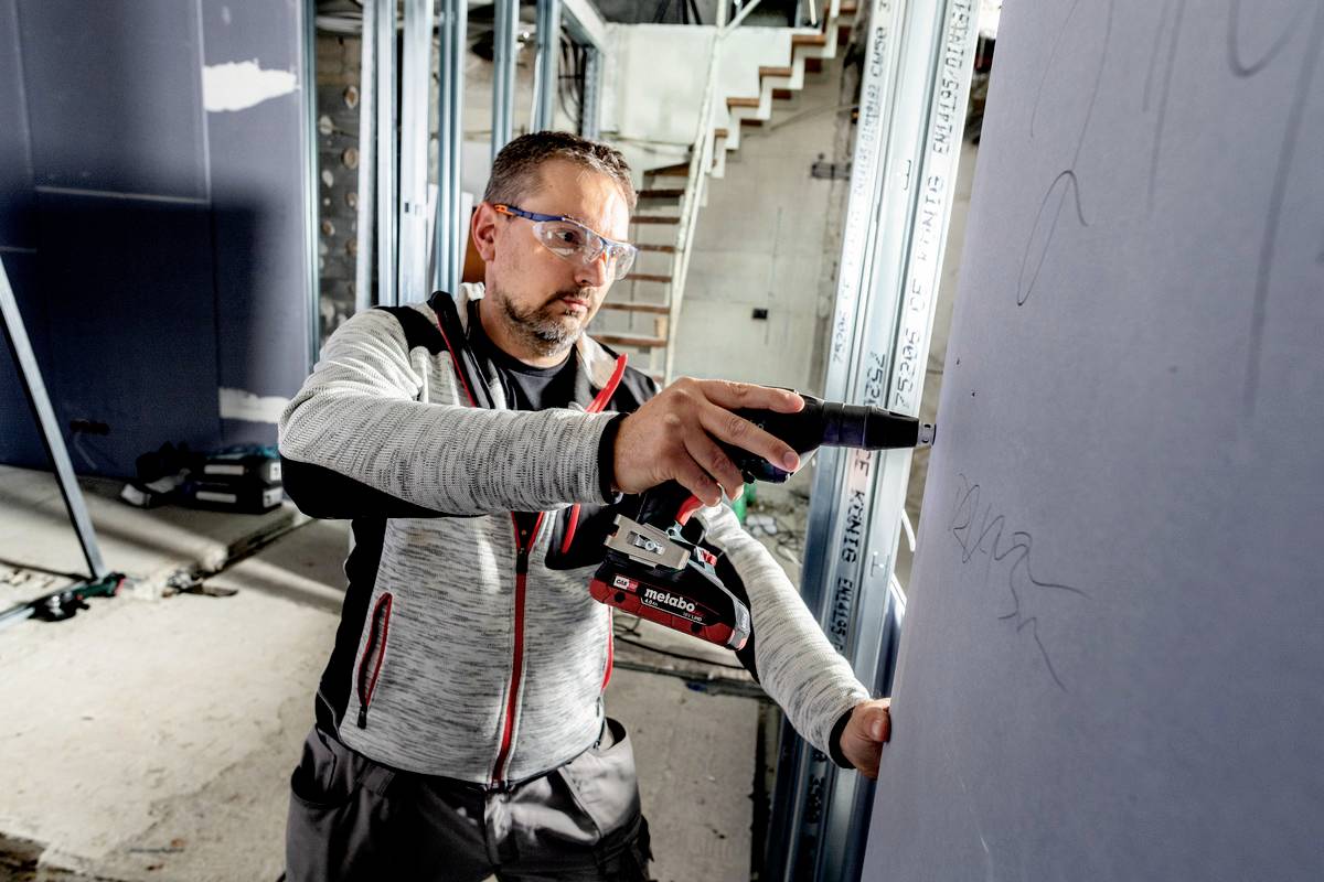 Ein Mann in Schutzbrille montiert eine Trockenbauwand mit einer Bohrmaschine in einem unfertigen Raum mit Metallrahmen.