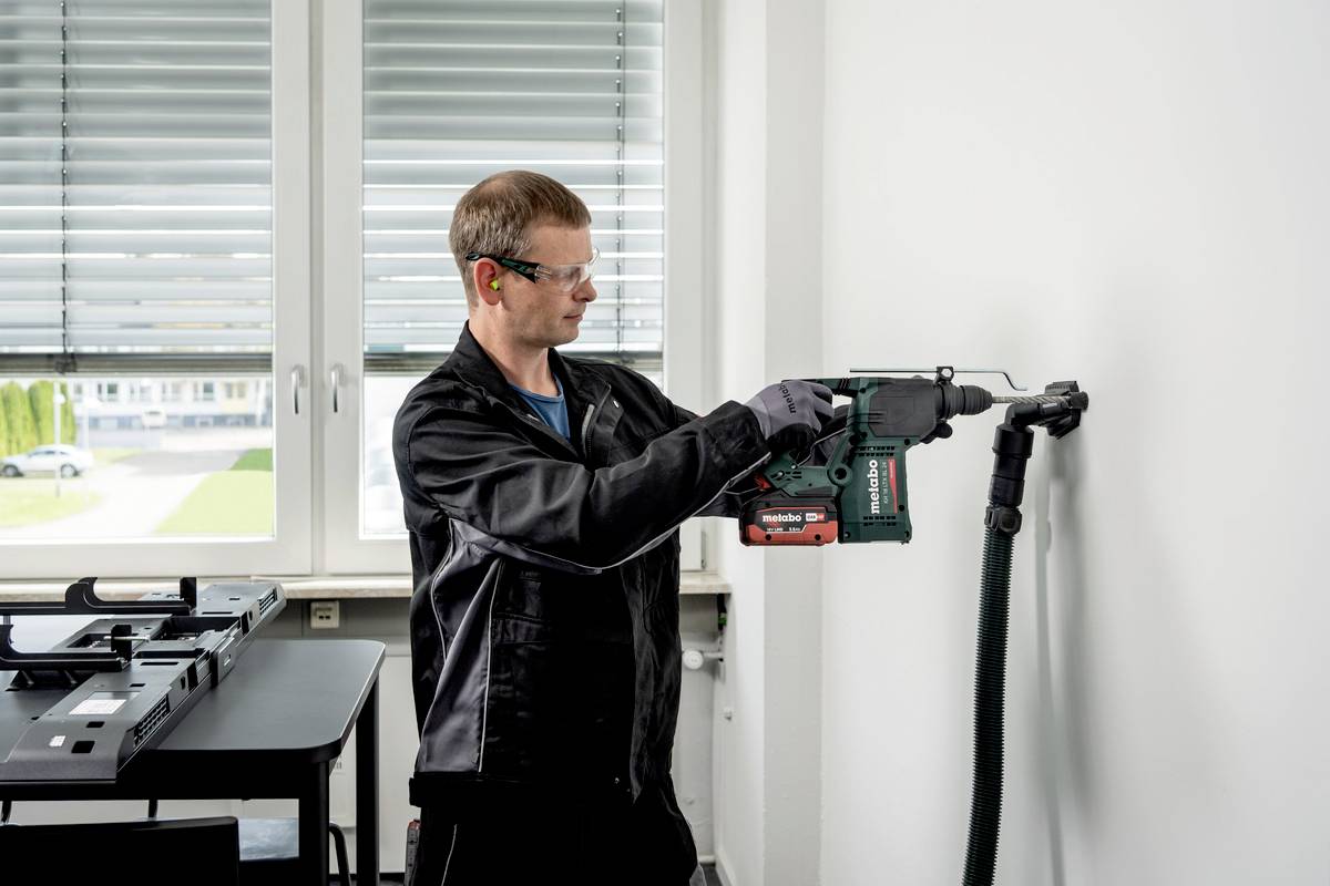 Ein Mann in Schutzbrille und Ohrschutz bohrt mit einer grünen Bohrmaschine in eine weiße Wand. Im Hintergrund sind Fenster zu sehen.