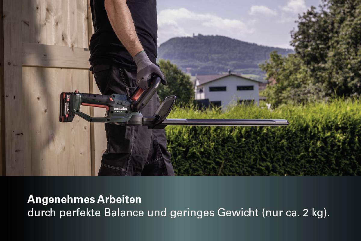 Person schneidet mit einem elektrischen Trimmer Hecken. Im Hintergrund sind ein Holzschuppen, Büsche und ein bewaldeter Hügel sichtbar.