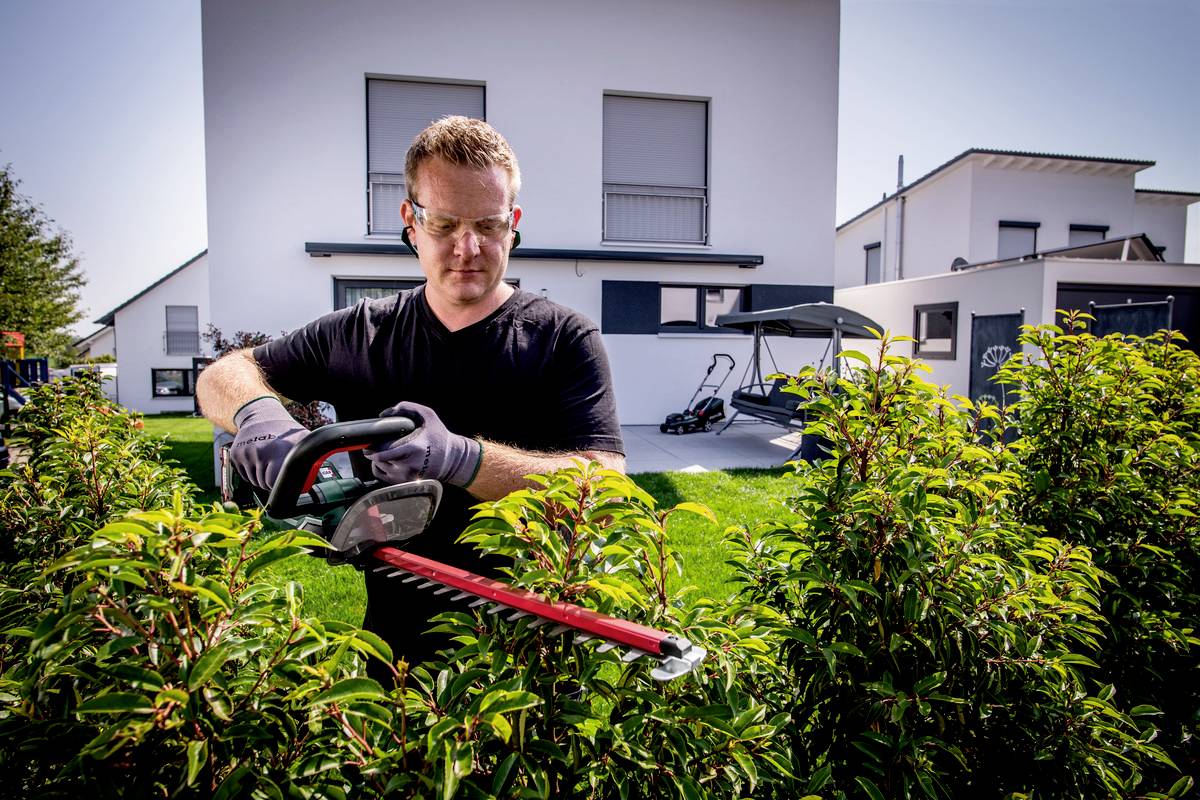 Ein Mann trimmt mit einer elektrischen Heckenschere die Hecke im Garten vor einem modernen weißen Haus bei sonnigem Wetter.
