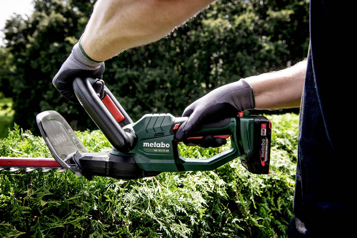 Eine Person schneidet eine Hecke mit einer elektrischen Heckenschere in einem Garten. Sie trägt Arbeitshandschuhe.