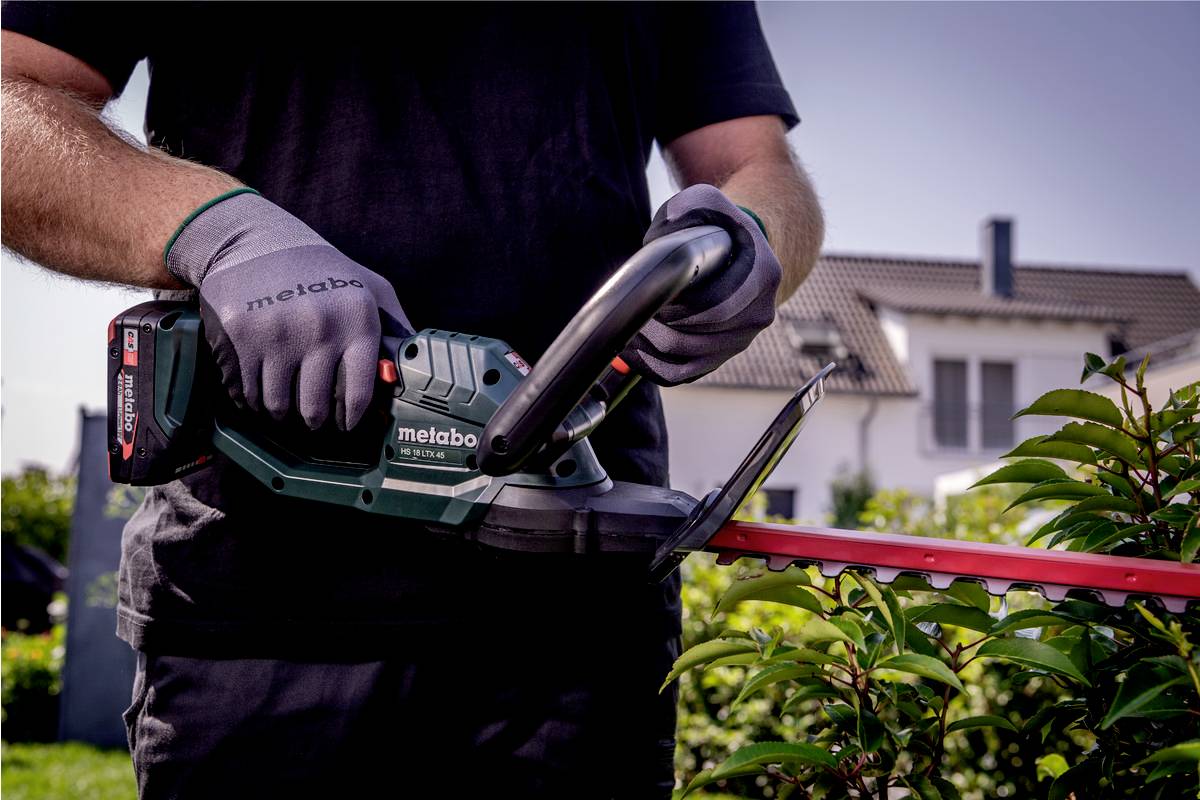 Eine Person bedient eine Metabo-Heckenschere, während sie Büsche im Garten trimmt. Im Hintergrund ist ein Wohnhaus zu sehen.