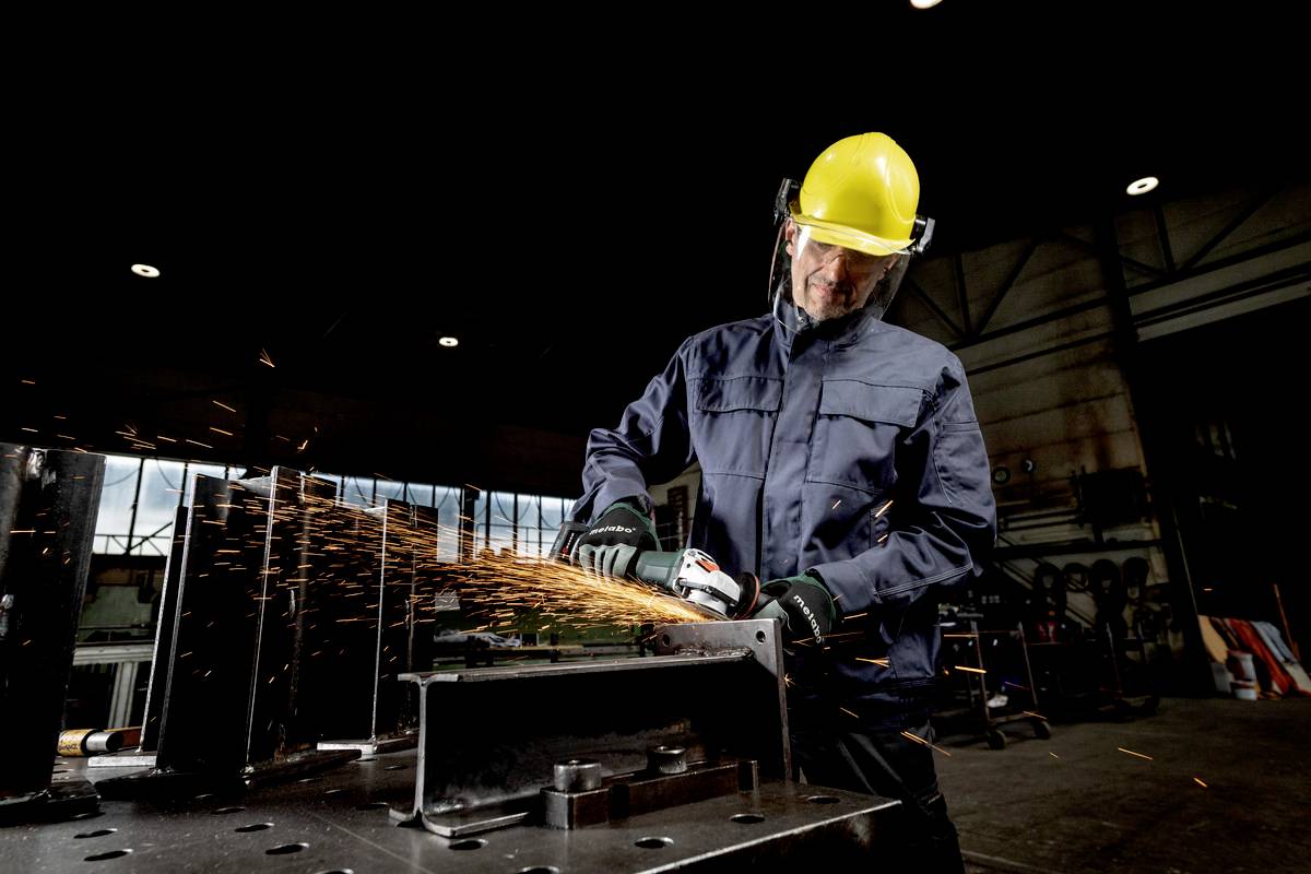 Ein Arbeiter mit gelbem Schutzhelm schleift Metall, Funken fliegen. Er trägt Schutzkleidung in einer Industriehalle.