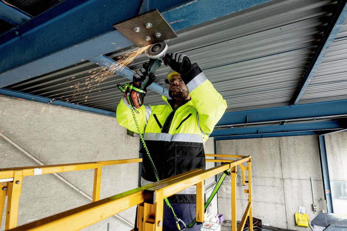 Eine Person arbeitet mit einem Winkelschleifer in einer metallischen Struktur auf einem Gerüst. Funken fliegen vom Bearbeitungspunkt.