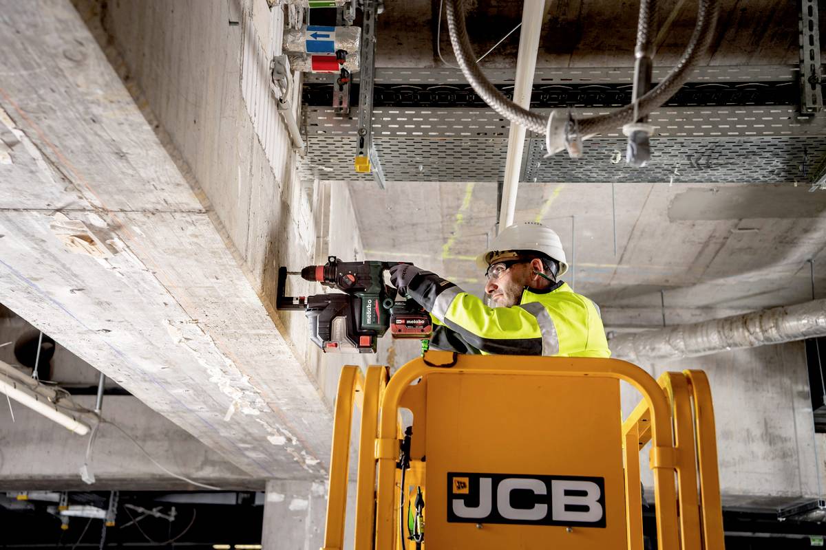 Ein Bauarbeiter steht auf einem gelben Hubsteiger, trägt Schutzkleidung und Helm, und bohrt mit einer Maschine in eine Betondecke.