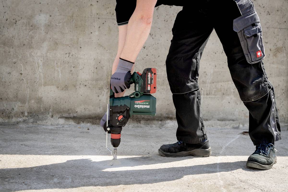 Eine Person in Arbeitskleidung benutzt einen Bohrhammer auf einem Betonboden. Hände in Schutzhandschuhen, Werkzeug vibriert.