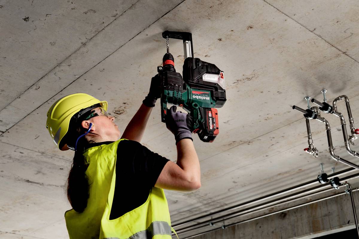 Arbeiterin mit gelber Helm und Schutzweste bohrt in Deckenplatte mit Akkuschrauber. Im Hintergrund sind Rohrleitungen zu sehen.