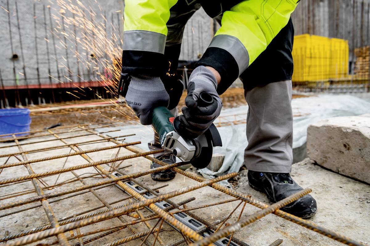 Eine Person schneidet Metallgitter mit einem Winkelschleifer auf einer Baustelle. Funken fliegen von der Klinge weg.