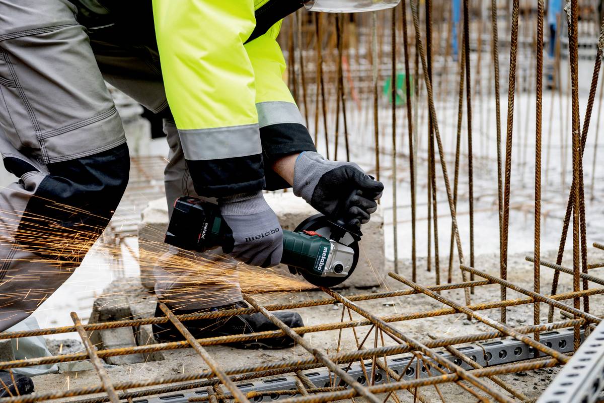 Bauarbeiter schneidet Stahlstangen mit einem Winkelschleifer auf einer Baustelle. Funken fliegen von der Maschine. Arbeiter trägt Sicherheitskleidung.