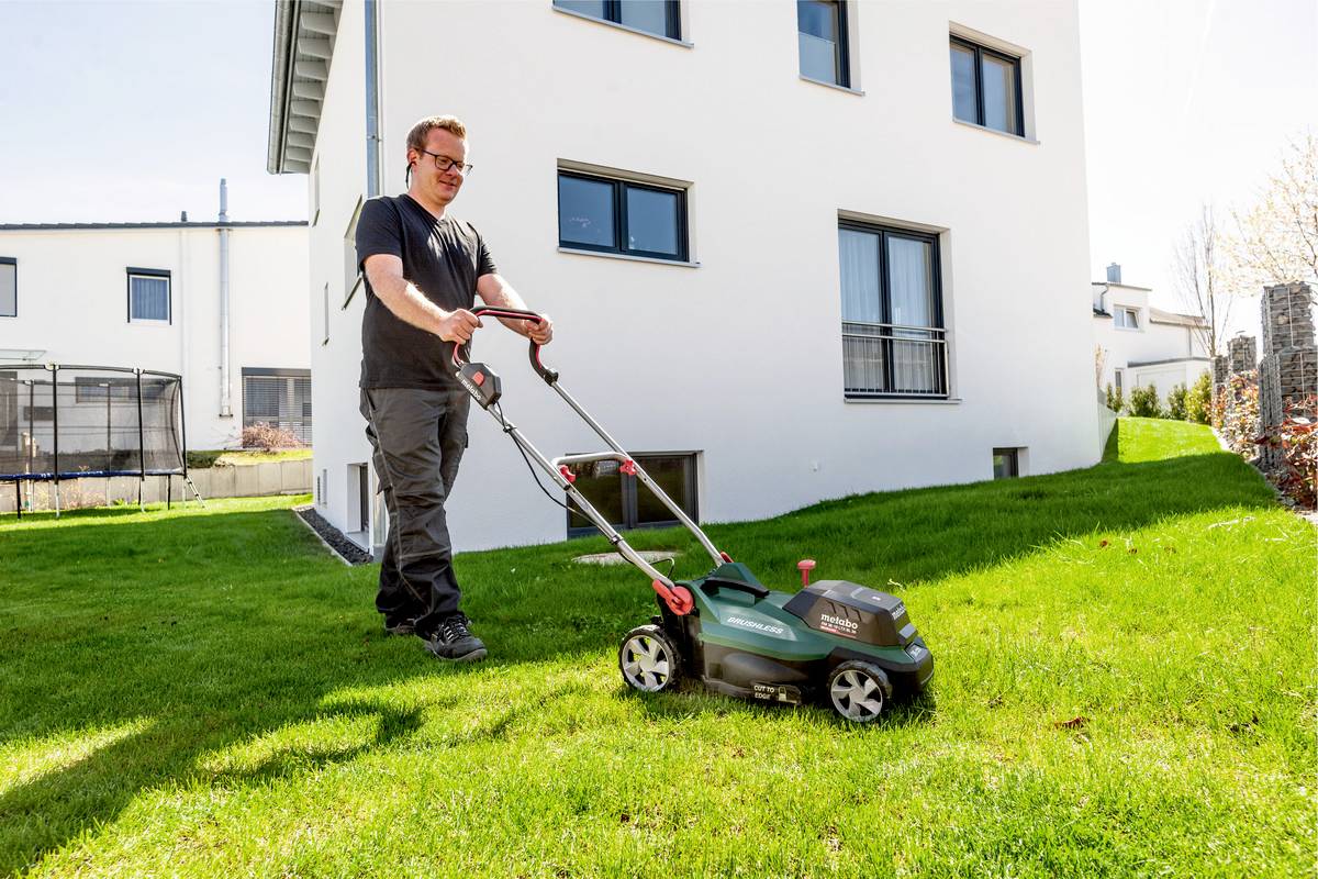 Ein Mann mäht den Rasen mit einem elektrischen Rasenmäher vor einem modernen, weißen Wohnhaus bei sonnigem Wetter.