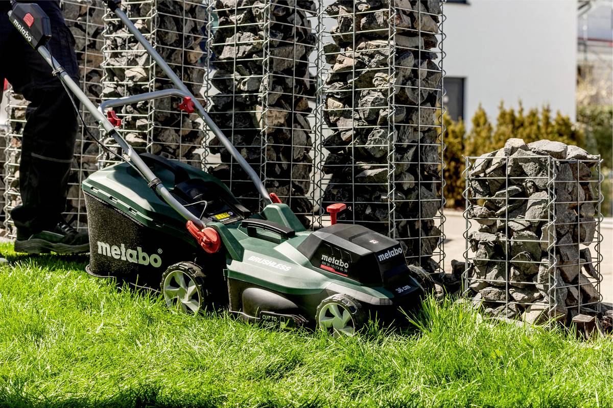 Eine Person mäht den Rasen mit einem Metabo-Rasenmäher neben mit Steinen gefüllten Gabionen in einem Garten.