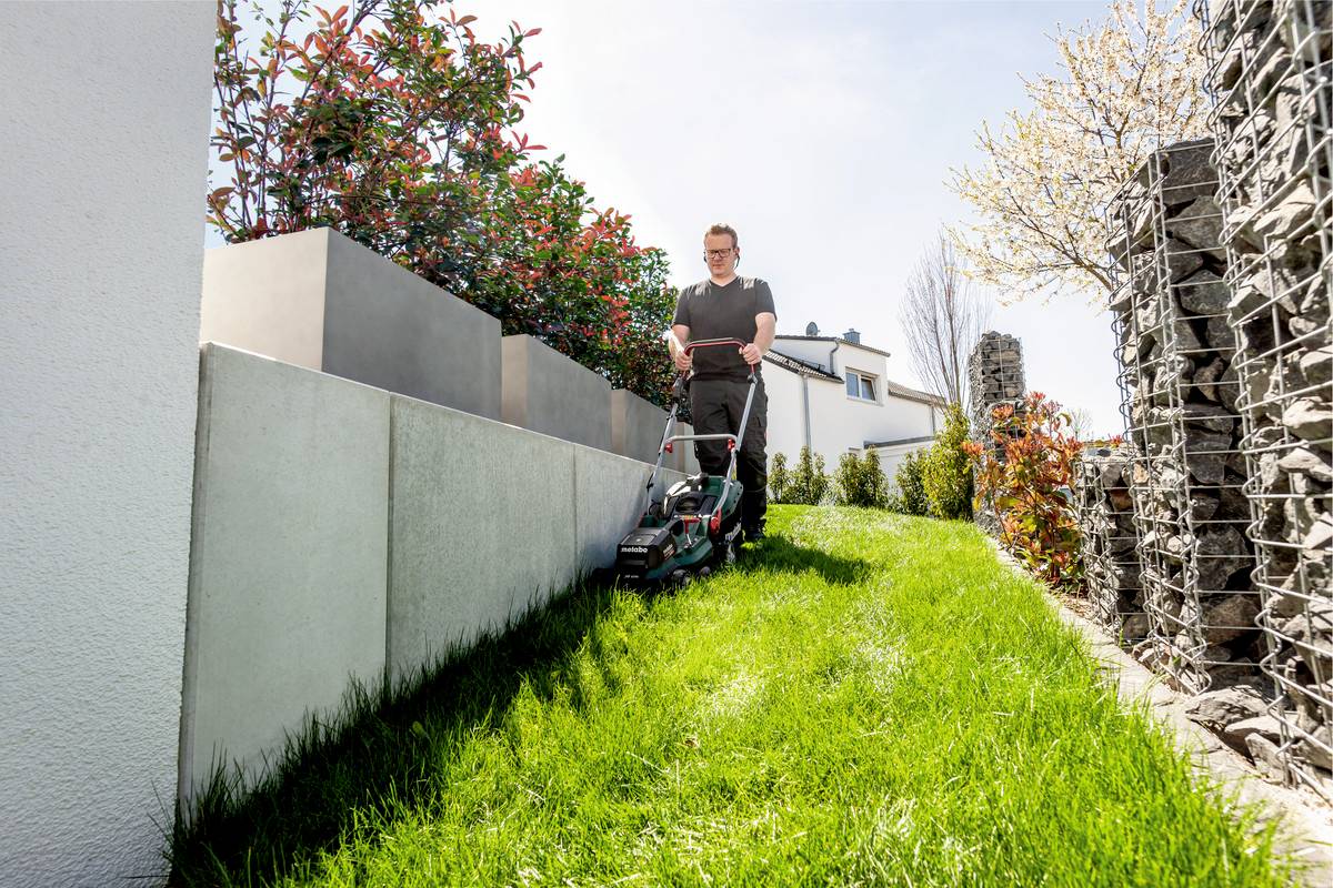 Eine Person mäht bei sonnigem Wetter den Rasen in einem modernen Garten mit weißen Pflanzkübeln und grünen Büschen.