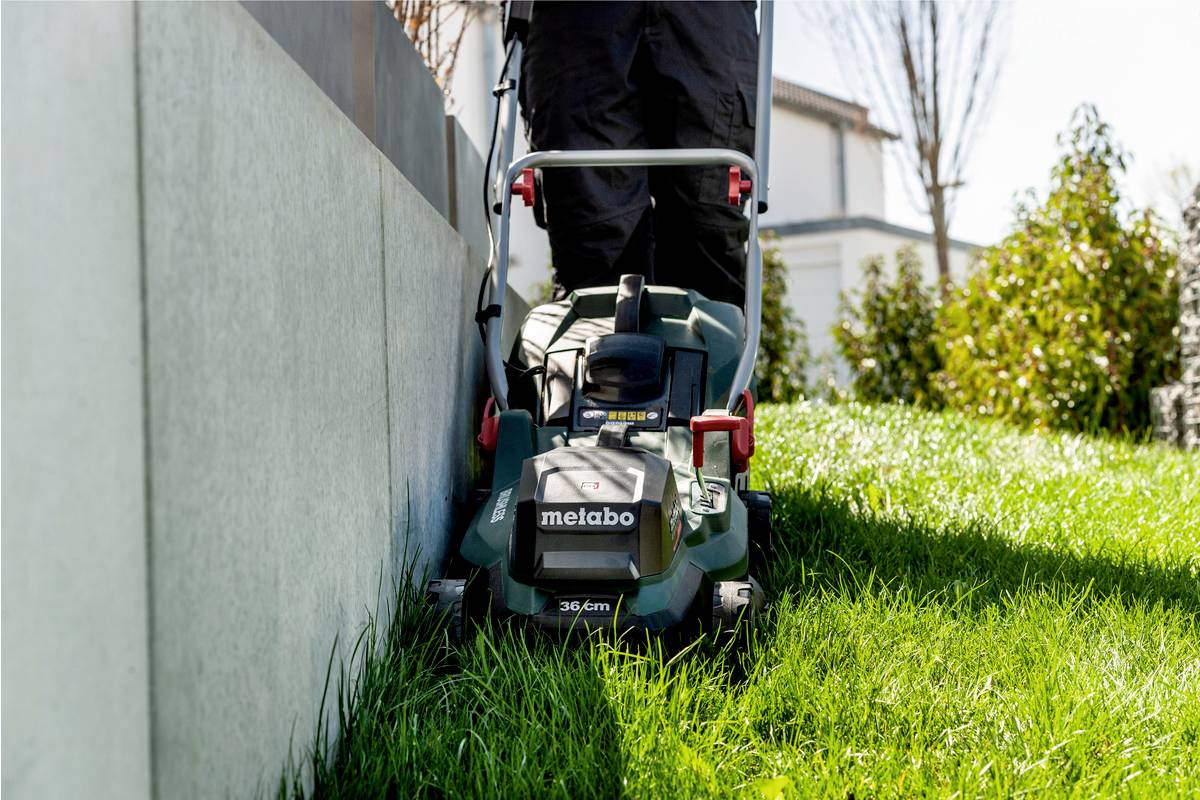 Eine Person benutzt einen Rasenmäher, um das Gras entlang eines betonierten Weges in einem Garten zu schneiden.