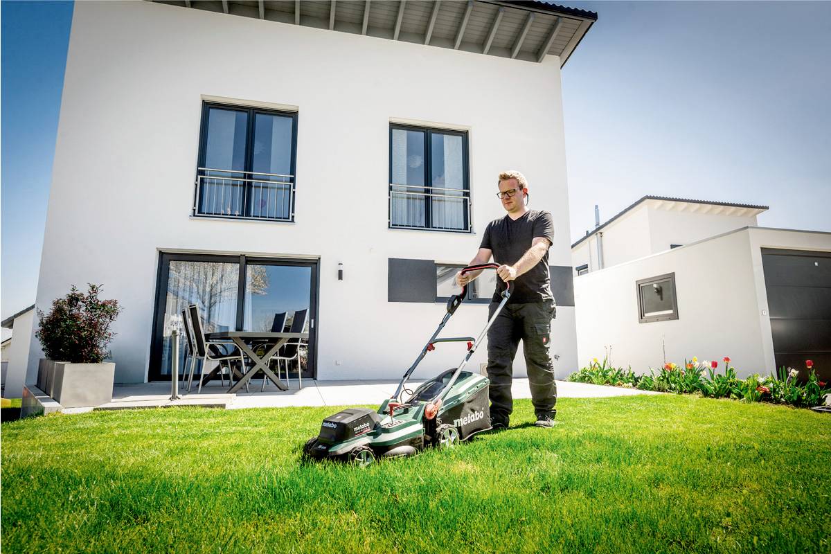 Eine Person mäht mit einem elektrischen Rasenmäher den Rasen vor einem modernen weißen Haus mit Balkon und Terrasse.