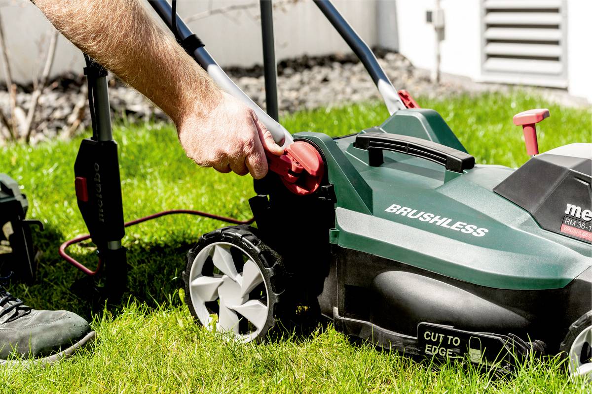 Person zieht den Stecker eines elektrischen Rasenmähers im Garten. Der Mäher ist grün und hat große, silberne Räder.