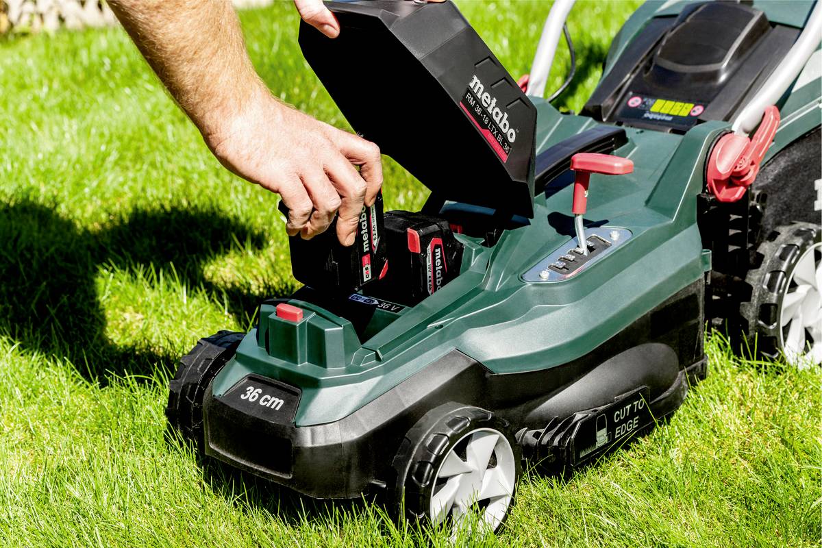 Eine Person wechselt den Akku eines grünen Rasenmähers im Garten. Der Mäher steht auf frischem, grünem Gras.