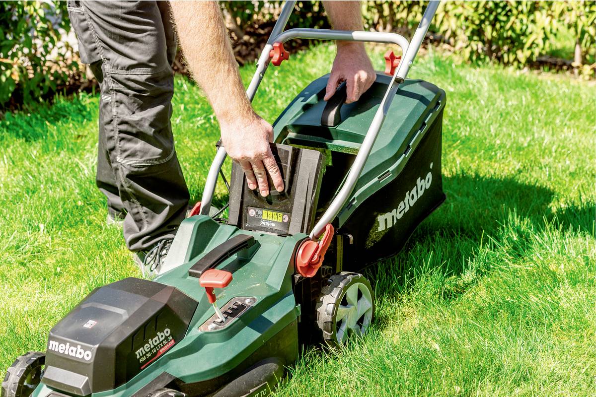 Eine Person wechselt den Akku eines grünen, batteriebetriebenen Rasenmähers im Garten. Der Rasenmäher steht auf grünem Gras.