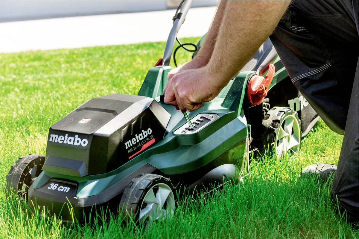 Eine Person justiert einen grünen Rasenmäher auf einer Grasfläche. Der Rasenmäher hat das Logo 'metabo' und die Angabe '36 cm'.