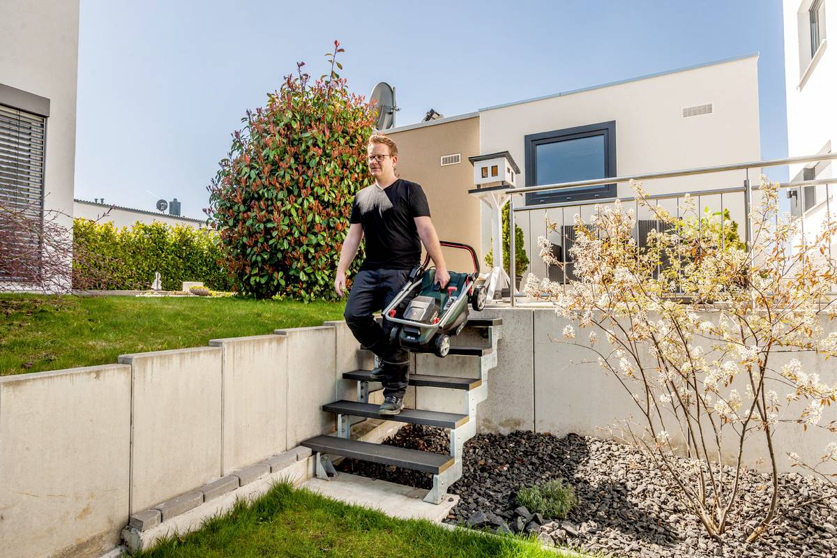 Eine Person trägt einen Rasenmäher die Treppe im Garten hinunter. Umgeben von Häusern und Pflanzen bei sonnigem Wetter.