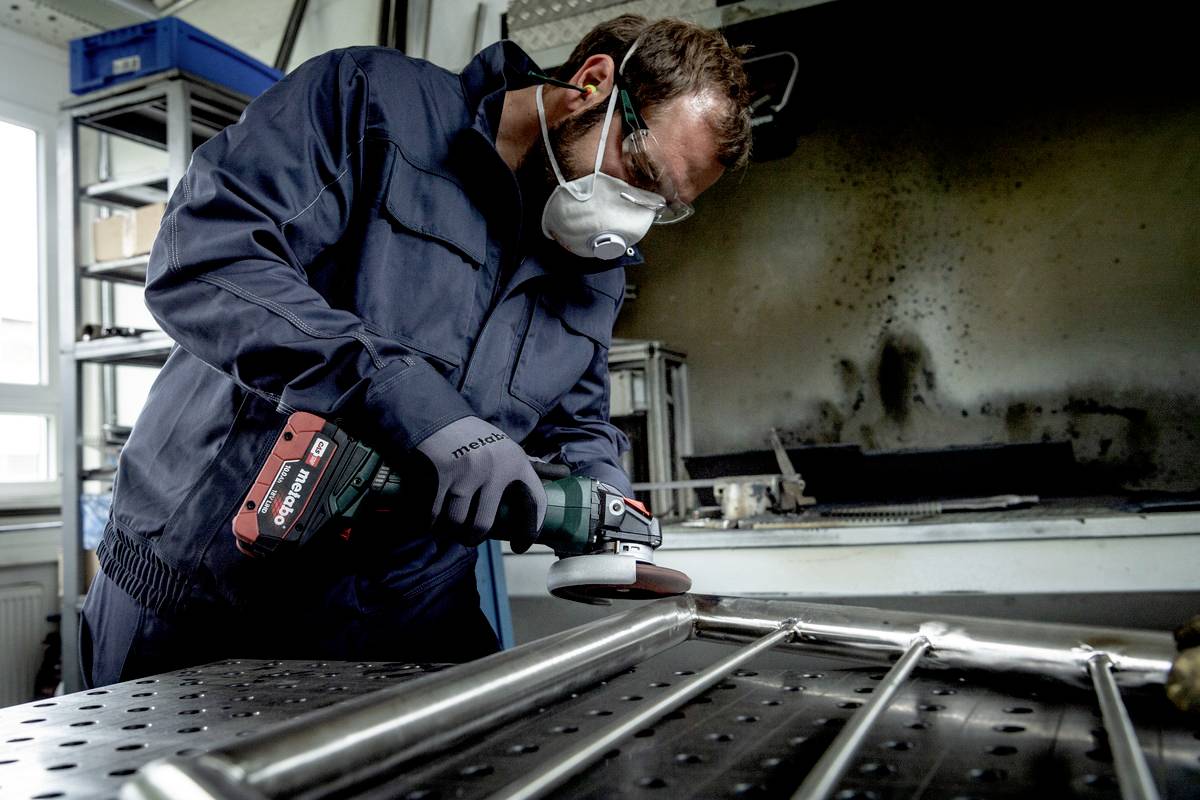 Ein Arbeiter in Schutzkleidung und -maske schleift ein Metallgestell in einer Werkstatt.